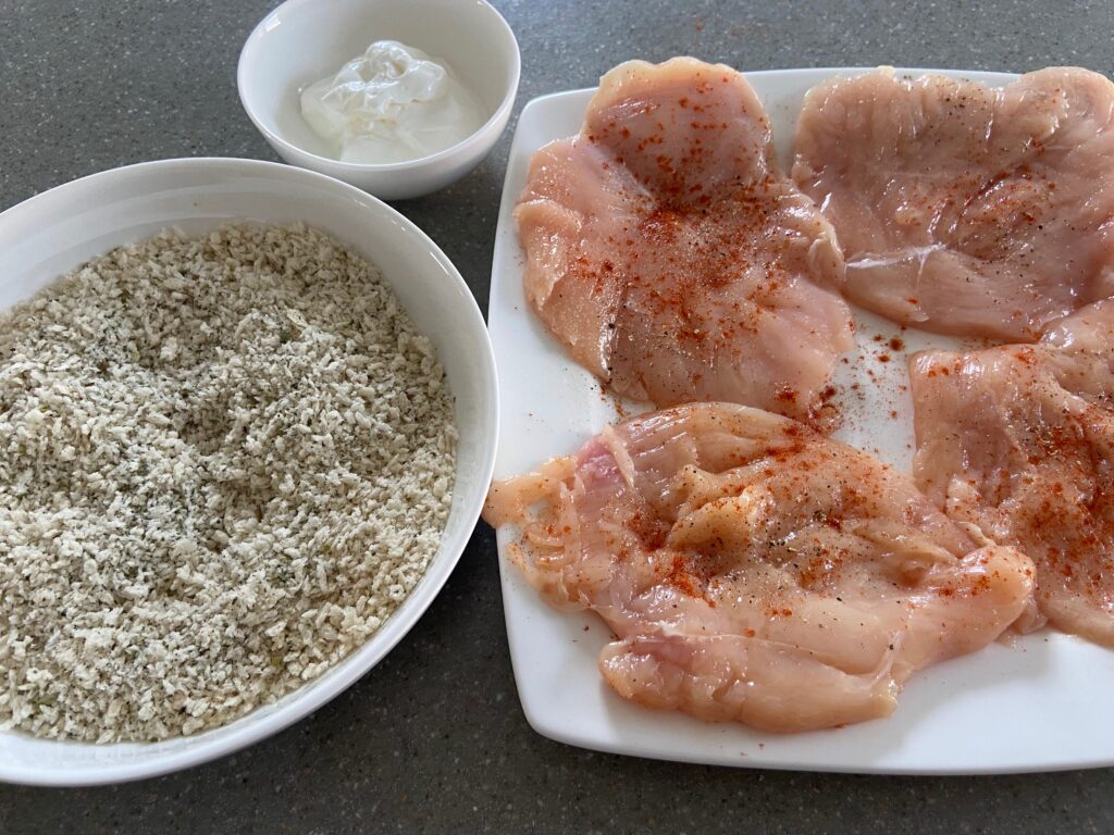 seasoned chicken cutlets and next to them is a bowl of Italian breadcrumbs and sour cream