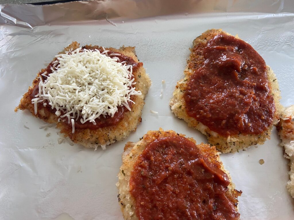 Chicken cutlets added to a foiled lined baking sheet and covered with tomato sauce and shredded cheese