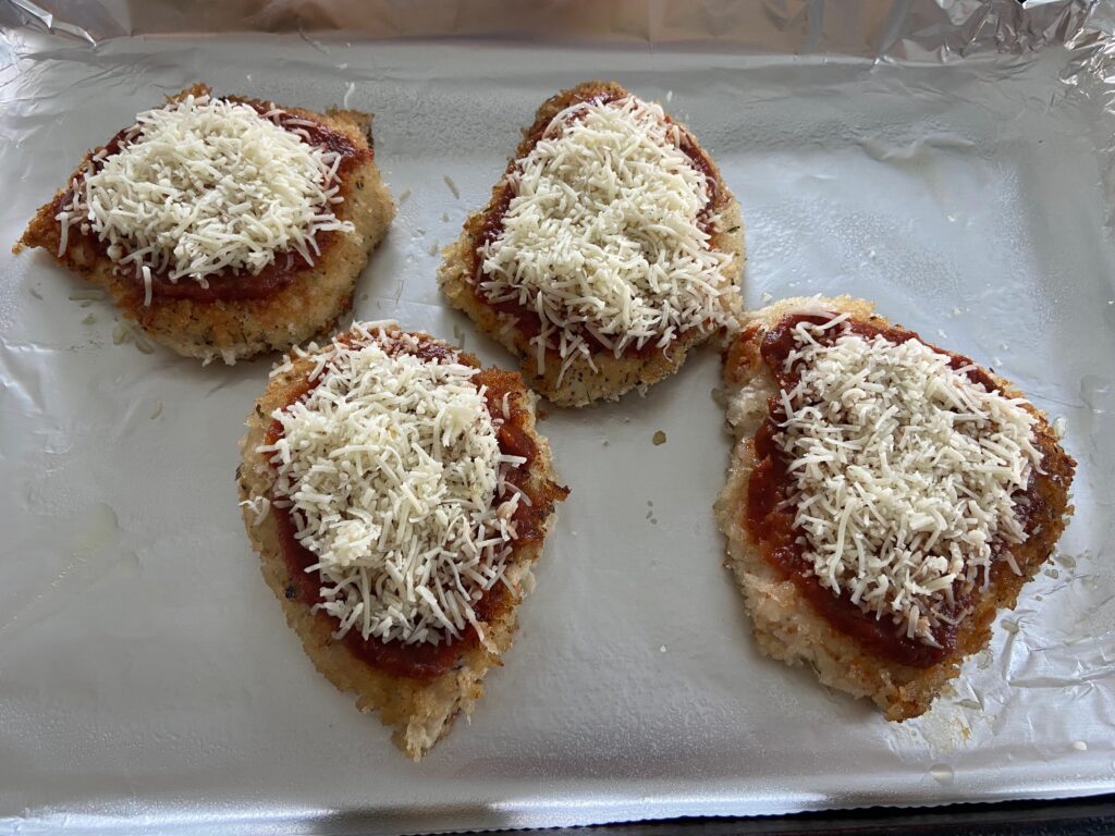 Chicken cutlets added to a foiled lined baking sheet and covered with tomato sauce and shredded cheese