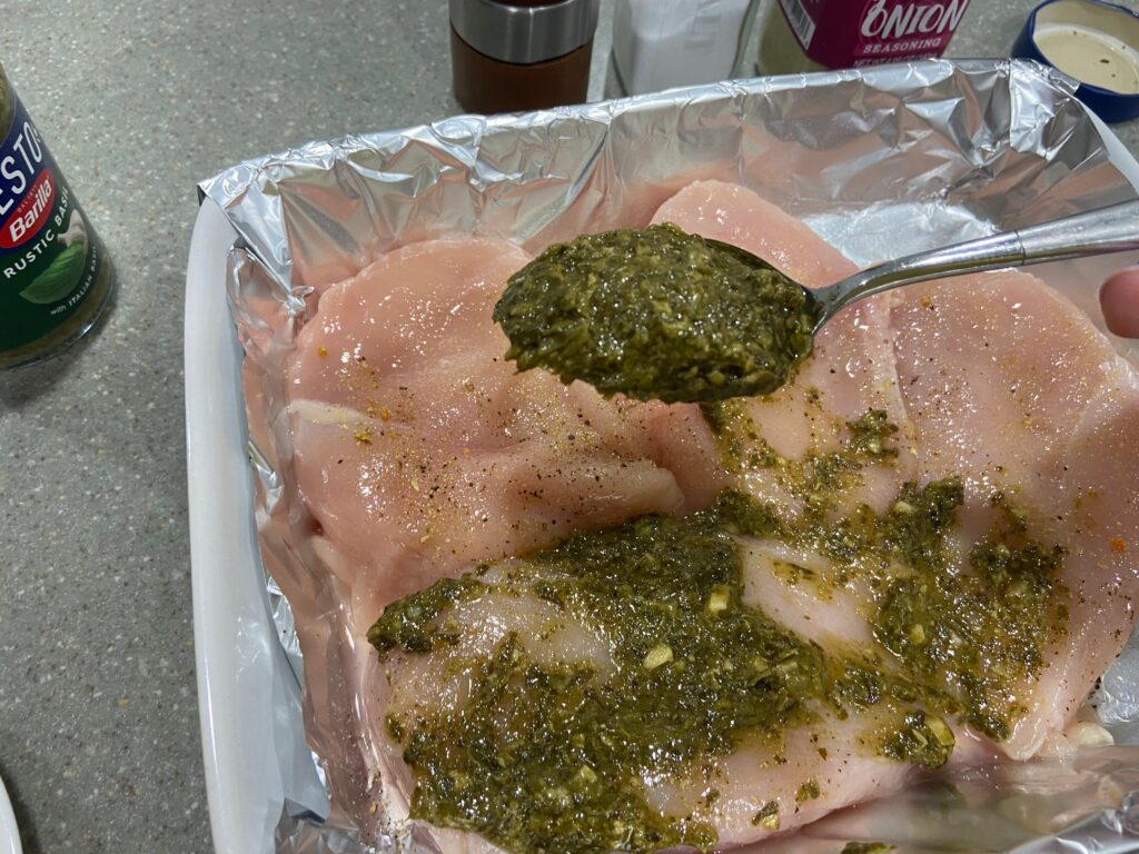 seasoned chicken breast cutlets arranged in a baking dish and a basil pesto spooned over the chicken