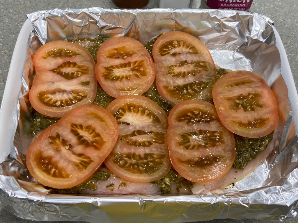 tomatoes slices on top of pesto spread out on top of seasoned chicken breast cutlets arranged in a baking dish