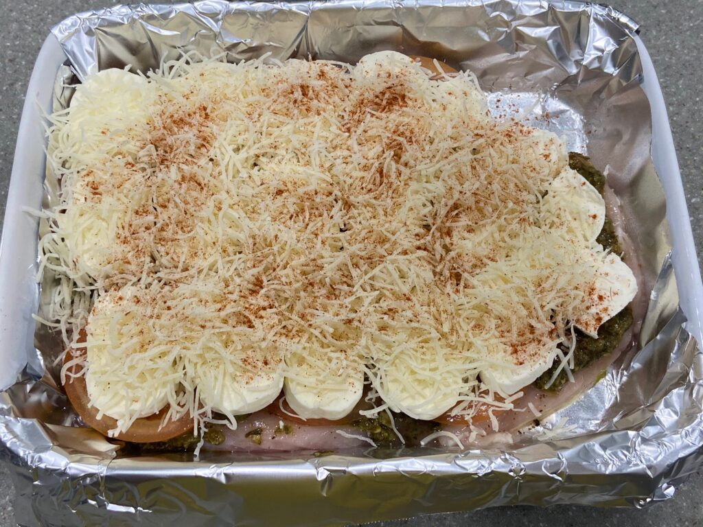 shredded cheese sprinkled on top of pesto spread out on top of seasoned chicken breast cutlets arranged in a baking dish